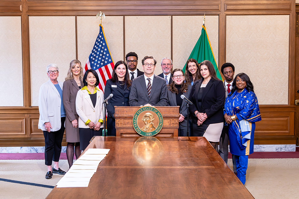 Photo of Meegan Brooks (in front of the green flag) is shown at the signing of HB 2274 by Washington Gov. Bob Ferguson, with the Washington Retail Association’s Interim Executive Director Alesha Shemwell (fourth from left) and Director of Policy and Government Affairs Crystal Leatherman (also in front of the green flag).